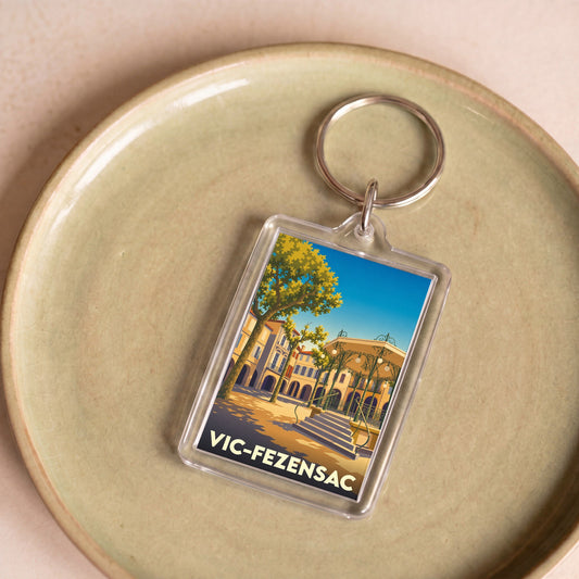 Porte-clés souvenir de la ville de Vic-Fezensac avec une illustration stylisée d'un kiosque et d'une place ensoleillée.