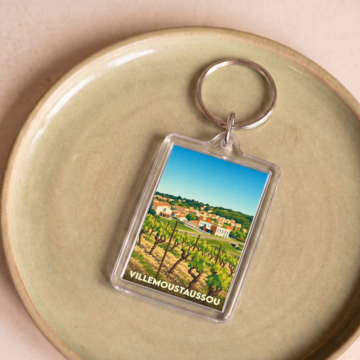 Porte-clés souvenir de la ville de Villemoustaussou avec une illustration stylisée de vignes et du village.