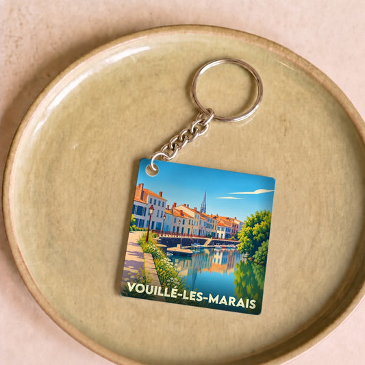 Porte-clés souvenir de la ville de Vouillé-les-Marais avec une illustration stylisée d'un canal bordé de maisons.