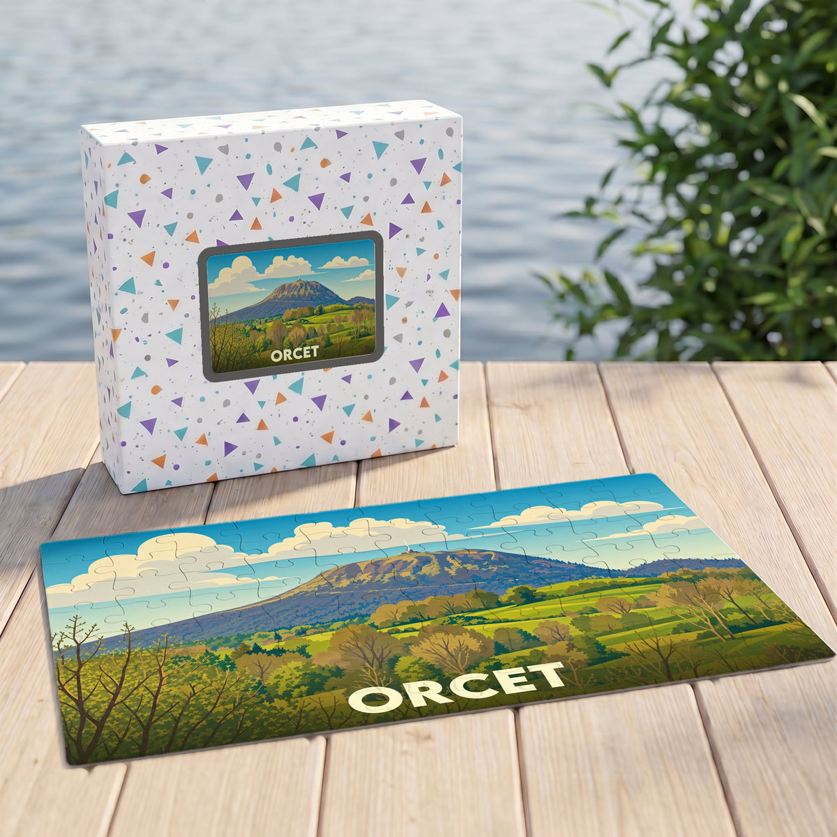 Puzzle souvenir de la ville d'Orcet avec son emballage, illustrant un paysage de collines verdoyantes sous un ciel azur.
