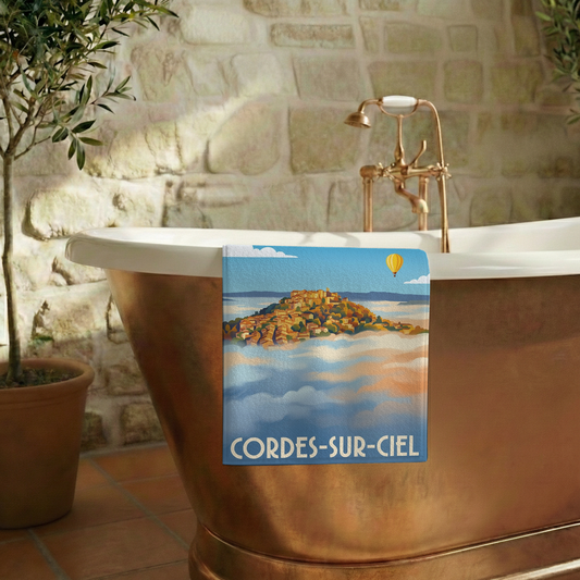Serviette de bain souvenir de Cordes-sur-Ciel avec une illustration stylisée de la cité émergeant des nuages.