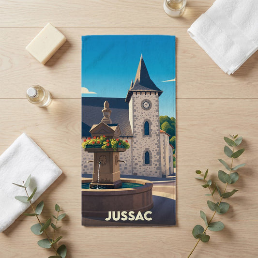 Serviette de bain souvenir de la ville de Jussac avec une illustration stylisée d'une fontaine et d'un clocher.