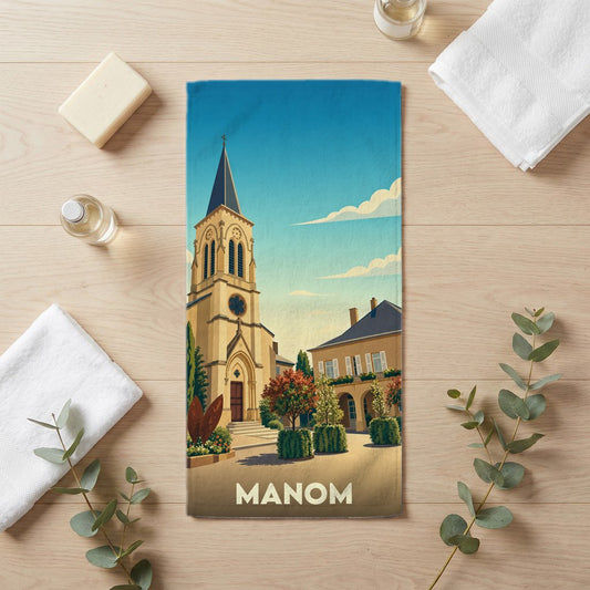 Serviette de bain souvenir de la ville de Manom avec une illustration stylisée d'une église et de maisons.