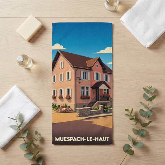 Serviette de bain souvenir de la ville de Muespach-le-Haut avec une illustration stylisée d'une maison alsacienne.