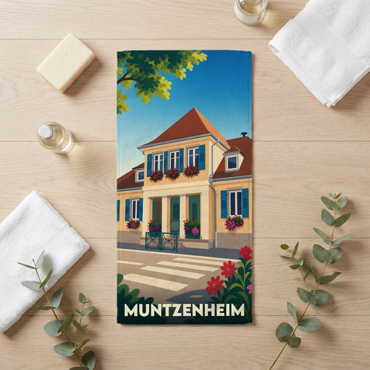 Serviette de bain souvenir de la ville de Muntzenheim avec une illustration stylisée d'un bâtiment traditionnel alsacien fleuri.
