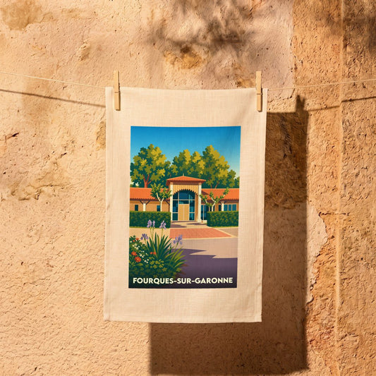 Torchon de cuisine arborant une illustration colorée et stylisée de Fourques-sur-Garonne, suspendu devant un mur en pierre.