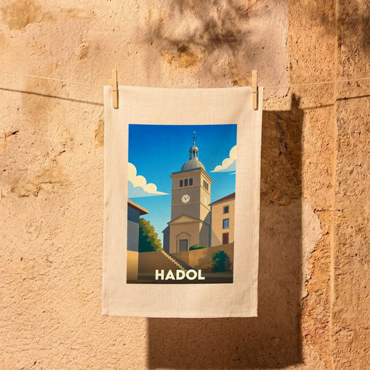 Torchon souvenir de la ville de Hadol illustré d'une église sous un ciel bleu, suspendu par des pinces à linge devant un mur en pierre.
