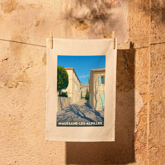 Torchon souvenir de la ville de Maussane-les-Alpilles avec une illustration stylisée d'une ruelle ensoleillée.