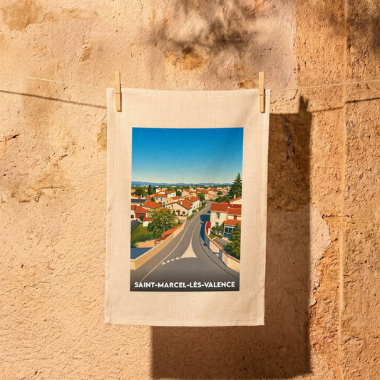 Torchon souvenir de Saint-Marcel-lès-Valence avec une illustration stylisée du village accroché à une corde à linge devant un mur en pierre.